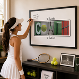 A woman in a white tennis outfit is writing a note on a framed tennis-themed art piece, which is mounted on a wall.