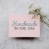 A pink card with the text "Handmade in the USA" written on it, accompanied by a small branch of white flowers.