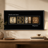 A framed artwork with the numbers "3" and "23" displayed in a creative way, along with the text "Sheila and Sam" and "Mignano" printed on it. The artwork is hanging on a wall above a wooden table, which has a vase and some books on it.