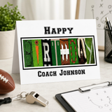A birthday card with the word "Happy" spelled out using football-related objects, placed on a desk with a football, clipboard, and other office supplies.