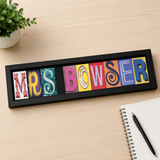 A framed sign displaying the name "Mrs. Bowser" in colorful letters is placed on a wooden surface next to a notebook and a pen.