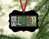 A black rectangular ornament with the word "IDEN" spelled out in various colors and shapes, hanging on a tree branch.