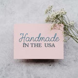 A pink card with the text "Handmade in the USA" written on it, accompanied by a small branch of white flowers.