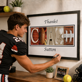 A young man in a football jersey is signing a framed letter that says "Thanks! Coach Sutton" on a wall.