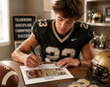 A young man in a black and gold football jersey is signing a paper with a pen, surrounded by football memorabilia, including a football helmet and a football.